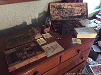 Wooden table displaying multiple vintage items including a dated wooden puzzle appear under a wooden box lid, visible ink stamps, metal safety pins, and brass bank.