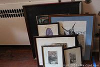 Framed artworks stacked against a wall. Visible prints include a bird print in wood frame with blue mat, various floral and scenic prints with white and black frames.