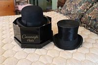 Two vintage gentleman's hats on a mattress: black velvet top hat and black bowler hat with Cavanagh Hats box.
