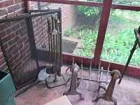 Photo showing the fireplace screen, andirons, log holder, and fireplace tools including a stand with tools like brush and poker, with a brick wall background.