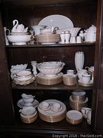 Cabinet shelves filled with white Lenox china pieces with gold rims showing assorted plates, bowls, cups, and decorative items.
