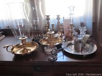Overview photo showing all brass and glass candle holders along with chimneys on a wooden surface in front of a window with sheer curtains.