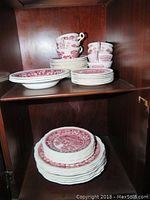 Full view of stacked plates, cups, bowls and saucers with pink floral and bridge transferware pattern inside wood cabinet.