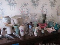 Display of various glass and porcelain vases, pitchers, and figurines including hobnail and Lenox pieces on wooden surface.