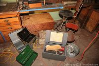 Wide view showing three metal tool boxes, U.S. Navy wooden boxes, vintage hand tools inside a gray metal box, metal buckets/strainers, and part of a wooden tool chest beneath a workbench.