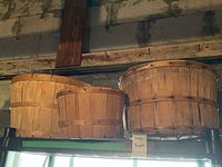 Main photo showing three wooden apple baskets on a shelf against a backdrop of an unfinished wall and ceiling with aged wood and some wiring visible.