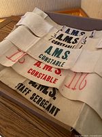 Seven cloth armbands folded in a box showing various printed ranks and A.M.S. lettering in different colors.