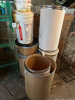 Seven assorted cardboard barrels and storage containers with metal rims, some with lids, displayed in a storage area.