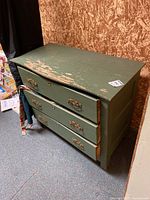 Green wooden dresser with three drawers. The dresser shows worn green paint with visible chipping, particularly on the top surface and drawer edges. Drawers feature decorative brass handles and keyhole escutcheons.
