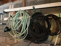 Photo showing assorted garden hoses including a light green hose with sprinkler attachment and several thick black rubber hoses, coiled and hanging on a wall.