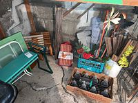 Overall view of lot with two green patio tables, kneeling bench, crate with painted vegetable garden stakes, wooden planks, and bottles in cardboard box