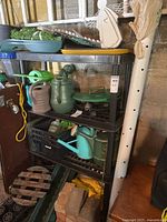 Shelf setup showing multiple plastic watering cans including green, beige, and turquoise colors, seedling pots, black plastic crates, and bricks at bottom shelf.