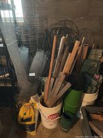 Photo showing assorted garden stakes bundled inside a white plastic bucket, several metal tomato cages, rolled metal wire fencing, decorative large metal trellises in background, and a partially visible bag of potting soil.