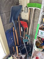 Photo showing assorted yard tools including shovel, broom, scoopers, crowbar, axe grouped together leaning against the wall