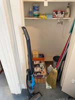 Photo shows Bissell vacuum standing with cleaning supplies on shelf including Clorox wipes, gloves, hand soap, broom, dusters, bucket and assorted lightbulbs packed in bins and boxes in small room or closet.