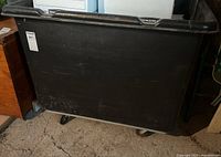 Front view of large black plastic storage bin on caster wheels, showing scratches and wear on the plastic surface.