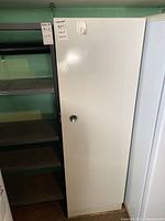 Tall white metal cabinet with handle and lock shown closed, next to shelves.