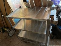 Full view of the three-tier metal hand cart showing metal shelves, tubular frame, and wheels. Signs of wear and use visible on surfaces.