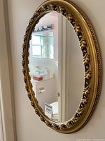 Full front view of the oval wall mirror showing the reflective surface and the ornate gold-tone plastic frame with decorative leaf motifs and beaded edge detail.