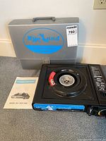 Photo showing the Myland portable gas stove, its hard carrying case with handle, and instruction manual placed on the floor.