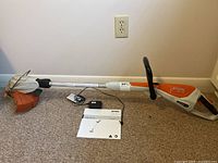 Full view of Stihl FSA 45 cordless weed wacker on carpet, laid flat with attached handle and trimming head visible.