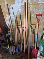 Photo of various yard tools including rakes, shovels, and a red plastic D-grip snow shovel against a wall