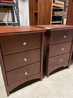 Two wooden bedside tables with three drawers each in a dark brown finish, showing the front view with metal knob handles.