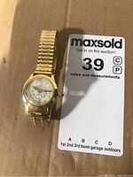 Gold-tone wristwatch with a Braille dial, clear cover, and gold folded link band shown against a cardboard background.