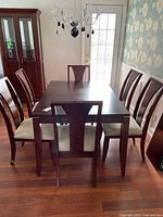Full view of rectangular wood dining table with six chairs arranged around it in a room with floral wallpaper and wood flooring.