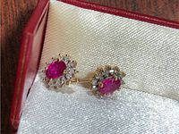 Pair displayed in red jewelry box showing oval ruby centers and diamond halos in yellow gold.