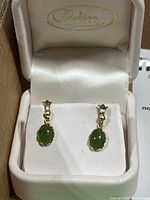 Pair of gold-tone drop earrings with oval polished green jade stones set in claw settings inside a cream-colored jewelry box.