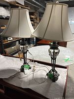 Pair of black and gold vintage style table lamps sitting on a wood surface with cream color fabric shades and visible cords.