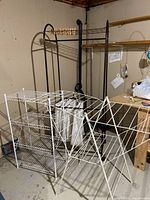 Three laundry racks: white folding drying rack, black laundry rack with hanging white cloth bags on wheels, and white mesh wire shelf unit.
