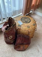 Photo showing the leather pouf with embroidery alongside two leather bucket bags on a carpeted floor near window curtains.