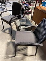 Two black resin wicker chairs with gray cushions and black wicker table with glass top shown from side angle in garage setting.