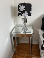 Full view of the glass mirrored side table with silver legs and decorative lamp on top near wall.