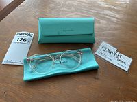 Clear round Tiffany & Co eyewear frames displayed with turquoise Tiffany branded leatherette carrying case and matching microfiber cleaning cloth on wooden surface.