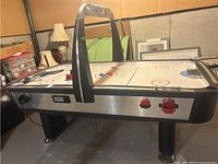 Photo showing the side and top view of the Sportcraft air hockey table with electronic scoreboard and red scoring knobs. The table stands on four black legs in a basement. Visible incomplete puck and glide on table surface.