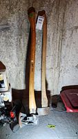 Two axes leaning against a wall including one with a metal head and wooden handle, and a separate metal tool at the base.