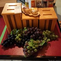Three wooden canisters with vertical dark stripes on a red surface with multiple bunches of faux grapes in front