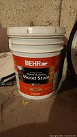 Front view of a large plastic pail of BEHR House & Fence Wood Stain with orange and black label and white lid.