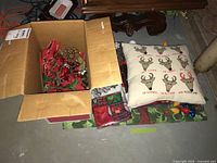 Box opened showing assorted Christmas decorations and holiday cushion with Christmas lights spread out on the ground