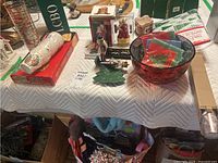 Holiday cups, coasters, figurines, red bowl, boxed Noma lights and other decorative items arranged on a table.