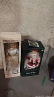 Two boxed Norm Motion-ette Christmas animated dolls, angel on left and Santa Claus on right, placed on concrete floor against wall.