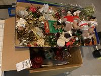 Box containing assorted Christmas decorations including plush reindeer ornaments, metallic ribbons, artificial leaves, pinecones, and baubles, showing overall worn condition.