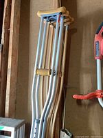 Photo showing both sets of crutches leaning against a wall, including two wooden crutches and two adjustable metal crutches with height adjustment holes and padded tops.