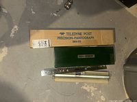 Photo showing original cardboard box labeled Teledyne Post Precision-Pantograph 38RA-010 with green hard case and metal rulers on floor