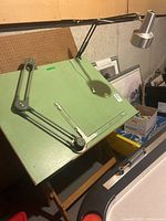 Drafting table featuring adjustable green slanted desktop with metal parallel straightedge and attached metal lamp, placed in basement setting.