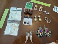 Overview photo showing the ten pairs of costume jewelry earrings arranged on a wooden surface, including three pairs new in package and various styles like pom-pom studs, pearl studs, and beaded dangles.