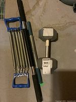 Photo of spring chest expander, dumbbell, hand grip strengthener, and spring arm exerciser on concrete floor.
